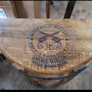K&ouml;nnte beinhalten: Ein halbkreisf&ouml;rmiges Fassdeckel-Regal aus Holz mit einem dunkelbraunen Aufdruck, der zwei gekreuzte Revolver sowie den Text SMOKE WAGON BOURBON, BIBAMUS MORIENDUM EST und SMOKE WAGON STRAIGHT BOURBON WHISKEY zeigt, befestigt mit einem schwarzen Metallband.
