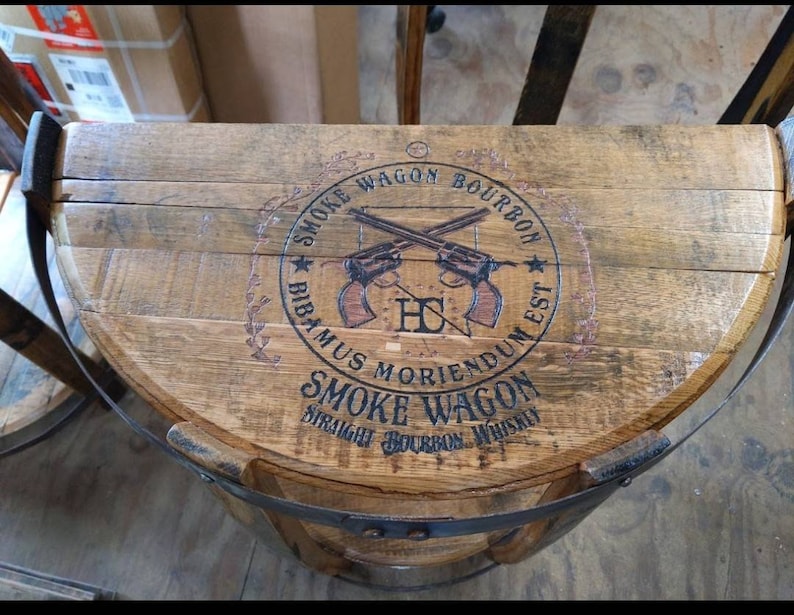 K&ouml;nnte beinhalten: Ein halbkreisf&ouml;rmiges Fassdeckel-Regal aus Holz mit einem dunkelbraunen Aufdruck, der zwei gekreuzte Revolver sowie den Text SMOKE WAGON BOURBON, BIBAMUS MORIENDUM EST und SMOKE WAGON STRAIGHT BOURBON WHISKEY zeigt, befestigt mit einem schwarzen Metallband.