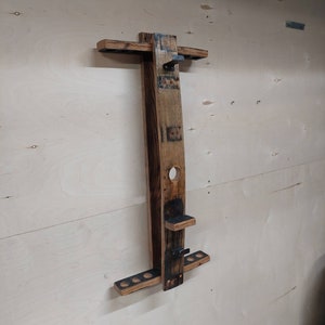 May include: A wooden wall-mounted rack made from a repurposed barrel stave, designed to hold pool cues. The rack features a top shelf for cue tips, a lower shelf for cue butts, and a metal hook for hanging a cue stick.