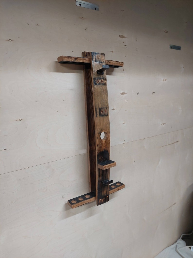 May include: A wooden wall-mounted rack made from a repurposed barrel stave, designed to hold pool cues. The rack features a top shelf for cue tips, a lower shelf for cue butts, and a metal hook for hanging a cue stick.