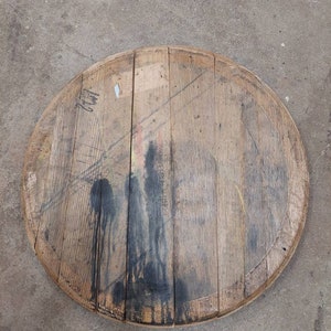 May include: A round wooden barrel top with a dark stain in the center. The wood is a light brown color and has a natural grain pattern. The top is slightly weathered and has a few small cracks.