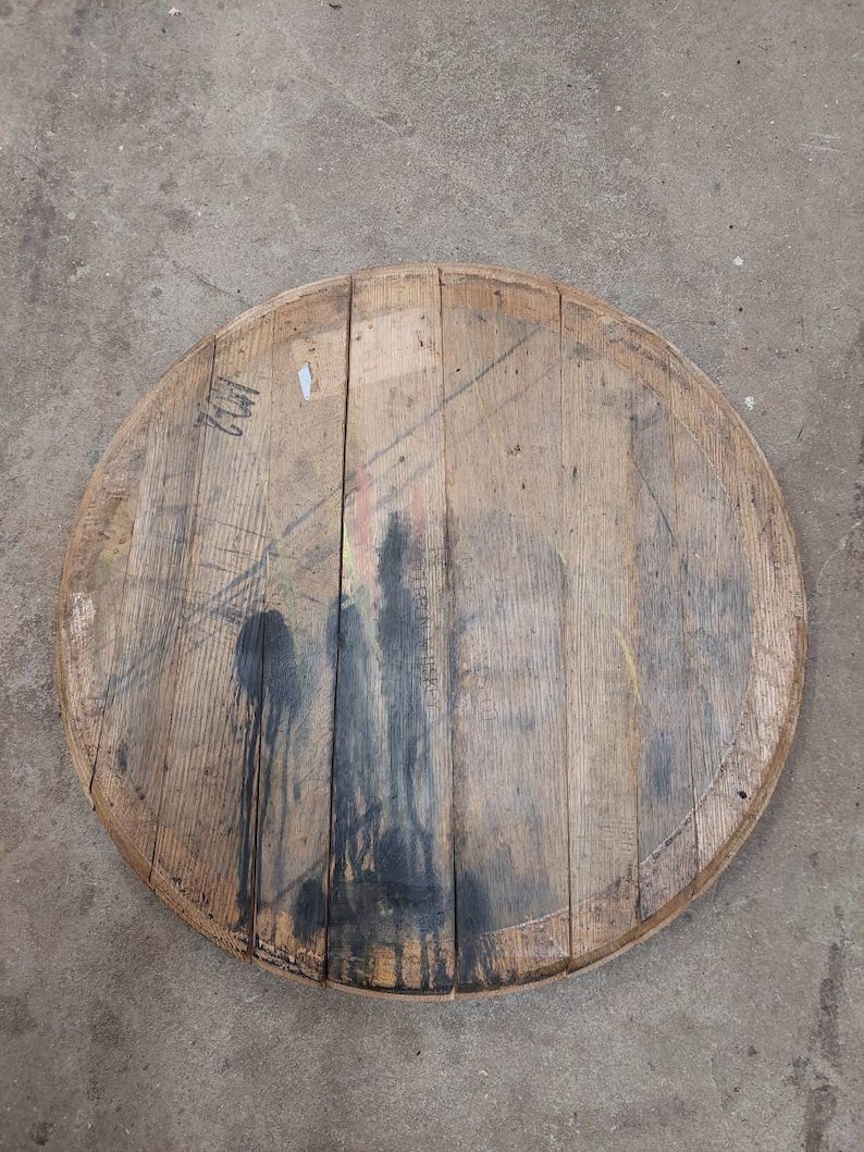 May include: A round wooden barrel top with a dark stain in the center. The wood is a light brown color and has a natural grain pattern. The top is slightly weathered and has a few small cracks.
