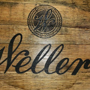 K&ouml;nnte beinhalten: Eine kreisf&ouml;rmige Holzoberfl&auml;che aus Fassdauben mit einem dunklen, eingebrannten Logo eines stilisierten Buchstabens W in einem kreisf&ouml;rmigen Kranz. Unter dem Logo steht das Wort Weller in einer gro&szlig;en, schwarzen Schreibschrift auf dem Holz.