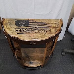 K&ouml;nnte beinhalten: Ein halbkreisf&ouml;rmiger Beistelltisch aus einem wiederverwendeten Holzfass mit einem dunklen Metallband und einer flachen Oberfl&auml;che. Das braun gebeizte Holz zeigt eine eingebrannte Grafik einer US-Flagge mit dem Jahr 1787 und dem Text We the People.