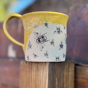 May include: A yellow ceramic mug with a white glaze and a black bee pattern. The mug has a rounded handle and a wide base.