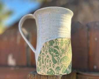 May include: White ceramic mug with a green mushroom pattern. The mug has a speckled glaze and a rounded handle.
