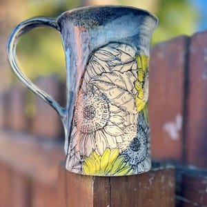 May include: A ceramic mug with a black and white sunflower design. The mug has a light blue glaze and a yellow rim.