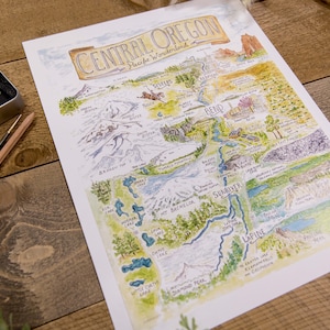 K&ouml;nnte beinhalten: Eine handgezeichnete Aquarellkarte von Central Oregon mit den Cascade Mountains, Fl&uuml;ssen, Seen und St&auml;dten. Die Karte tr&auml;gt den Titel "Central Oregon, Pacific Wonderland".
