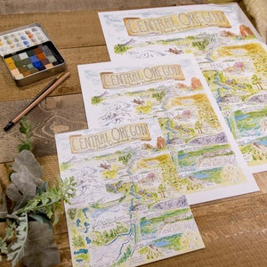 K&ouml;nnte beinhalten: Drei verschiedene Gr&ouml;&szlig;en von Karten von Central Oregon, gedruckt auf wei&szlig;em Papier, mit handgezeichneten Illustrationen der Sehensw&uuml;rdigkeiten und Naturmerkmale der Region. Die Karten liegen auf einer Holzoberfl&auml;che mit einem Aquarellfarbenset und Bleistiften.