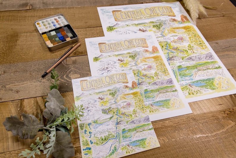 K&ouml;nnte beinhalten: Drei verschiedene Gr&ouml;&szlig;en von Karten von Central Oregon, gedruckt auf wei&szlig;em Papier, mit handgezeichneten Illustrationen der Sehensw&uuml;rdigkeiten und Naturmerkmale der Region. Die Karten liegen auf einer Holzoberfl&auml;che mit einem Aquarellfarbenset und Bleistiften.