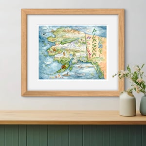 May include: A watercolor illustration of a map of Alaska with various animals and plants. The map is framed in a light wood frame and is hanging on a wall.