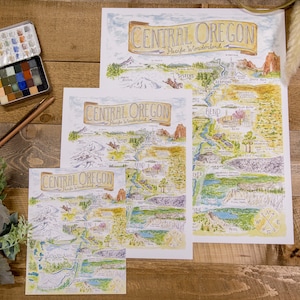 K&ouml;nnte beinhalten: Drei verschiedene Gr&ouml;&szlig;en von Karten der Central Oregon Pacific Wonderland, gedruckt auf wei&szlig;em Papier mit handgezeichneten Illustrationen im Aquarellstil. Die Karten zeigen einen Kompass, Fl&uuml;sse, Berge und andere Sehensw&uuml;rdigkeiten.