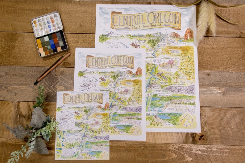 K&ouml;nnte beinhalten: Drei verschiedene Gr&ouml;&szlig;en von Karten der Central Oregon Pacific Wonderland, gedruckt auf wei&szlig;em Papier mit handgezeichneten Illustrationen im Aquarellstil. Die Karten zeigen einen Kompass, Fl&uuml;sse, Berge und andere Sehensw&uuml;rdigkeiten.