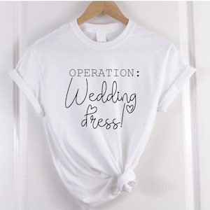 Pode incluir: Camiseta branca com texto preto que diz "OPERATION: Wedding Dress!" com um pequeno gráfico de coração.