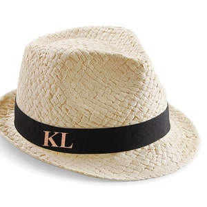 Peut inclure: Chapeau fedora en paille beige avec une texture tressée. Il est orné d'un ruban noir portant les initiales "KL" en couleur cuivre. Le chapeau est posé sur un fond blanc.