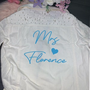 Könnte beinhalten: Weiße Jeansjacke mit Perlenbesatz an den Schultern. Auf der Rückseite der Jacke steht in blau "Mrs. Florence" mit einem blauen Herz.