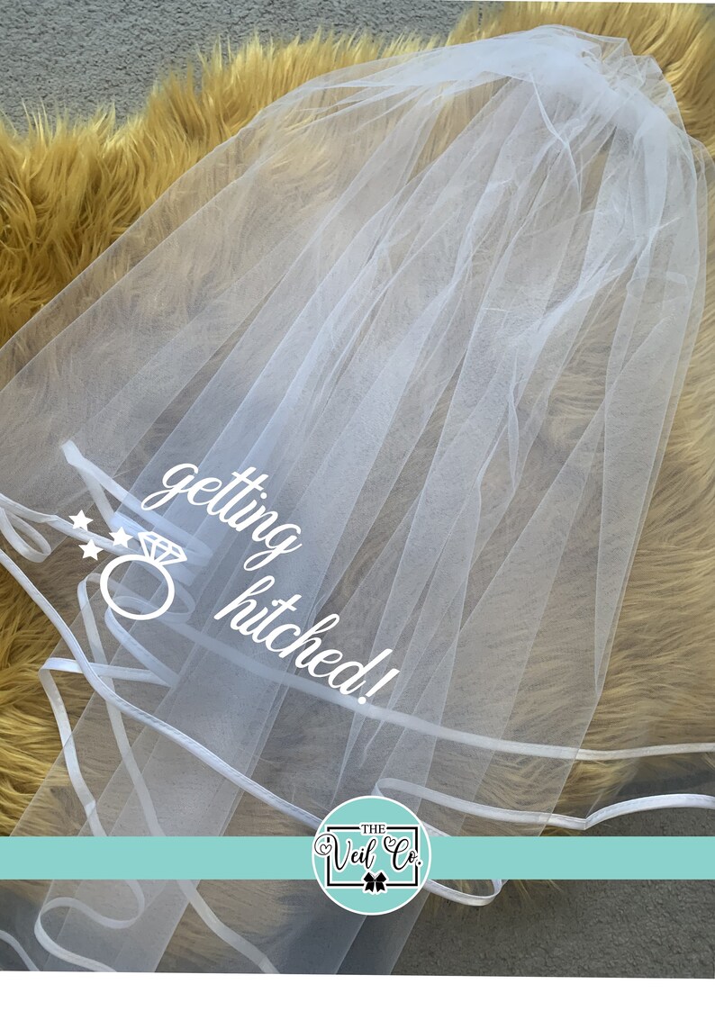 May include: White bridal veil with a white satin trim and the text "getting hitched!" with a diamond ring graphic. The veil is on a fuzzy yellow surface. The text "THE Veil Co." is in a circle with a bow.