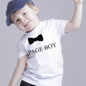 Op de afbeelding: Een jonge jongen draagt een wit T-shirt met een zwarte strik en de tekst "PAGE BOY". Hij draagt ook een blauwe baseballpet en een blauwe spijkerbroek.