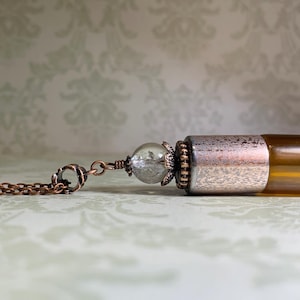 May include: A copper-toned chain attached to a clear, spherical glass bead with a decorative metal cap. The bead connects to a cylindrical amber-colored glass vial featuring a textured, metallic silver-toned cap at the top end of the container.