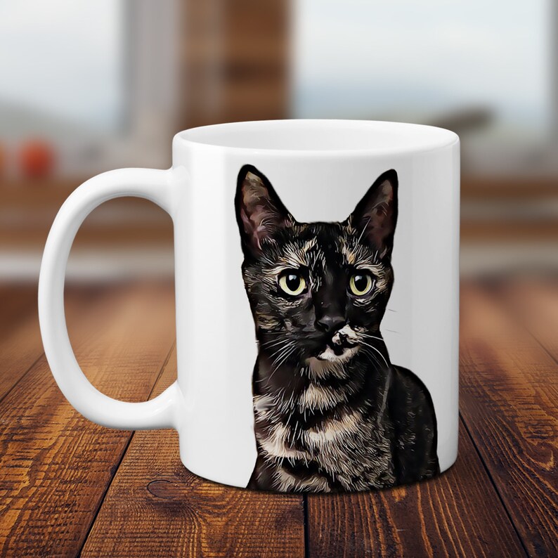 K&ouml;nnte beinhalten: Wei&szlig;e Keramiktasse mit einer Illustration einer schwarz-braunen Tabby-Katze. Die Katze hat gr&uuml;ne Augen und schaut direkt auf den Betrachter.