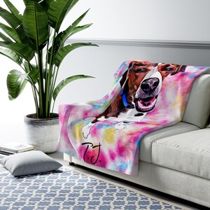 May include: A soft, colorful throw blanket featuring a watercolor-style image of a happy dog. The blanket has a tie-dye effect with pink, yellow, and blue hues. The dog is brown and white with a blue collar. The initials "T.J." are written on the blanket.