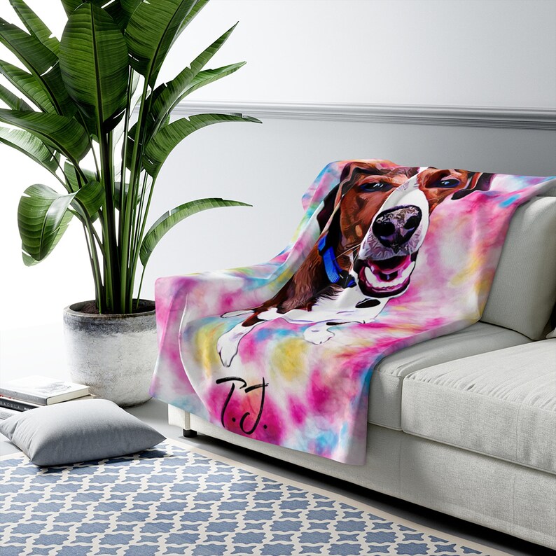 May include: A soft, colorful throw blanket featuring a watercolor-style image of a happy dog. The blanket has a tie-dye effect with pink, yellow, and blue hues. The dog is brown and white with a blue collar. The initials "T.J." are written on the blanket.