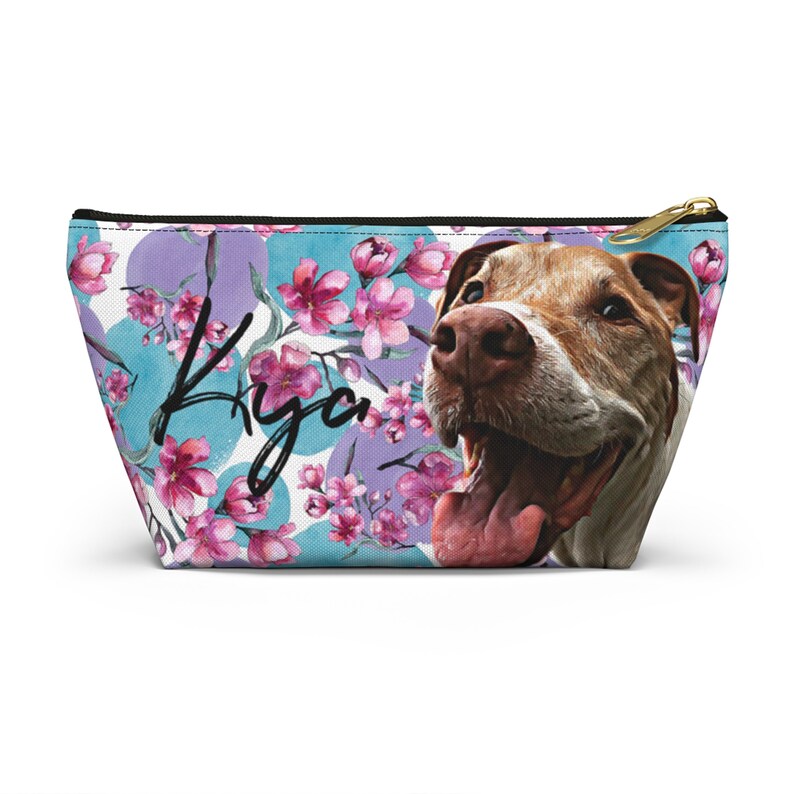 Puede incluir: Una bolsa blanca con cremallera con un estampado floral con flores rosas y c&iacute;rculos azules y morados. El nombre "Kya" est&aacute; escrito en letra negra en la bolsa. Se ve la cara de un perro marr&oacute;n y blanco en el lado derecho de la bolsa.
