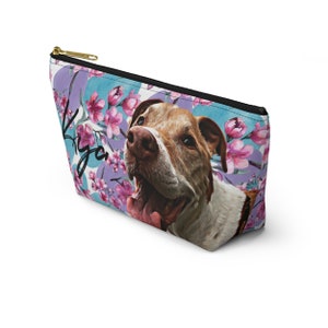 Puede incluir: Una bolsa de cosm&eacute;ticos con un patr&oacute;n floral de flores rosas y hojas moradas sobre un fondo azul. Un perro blanco y marr&oacute;n con la nariz rosa y la lengua es presentado en la bolsa. El nombre del perro, "Iya", est&aacute; escrito en cursiva negra en la bolsa.