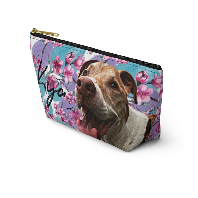 Puede incluir: Una bolsa de cosm&eacute;ticos con un patr&oacute;n floral de flores rosas y hojas moradas sobre un fondo azul. Un perro blanco y marr&oacute;n con la nariz rosa y la lengua es presentado en la bolsa. El nombre del perro, "Iya", est&aacute; escrito en cursiva negra en la bolsa.
