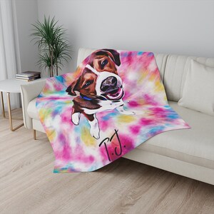 May include: A throw blanket featuring a colorful tie-dye background with a portrait of a dog. The dog is brown and white with a blue collar. The blanket has the handwritten initials "T.J." in black.