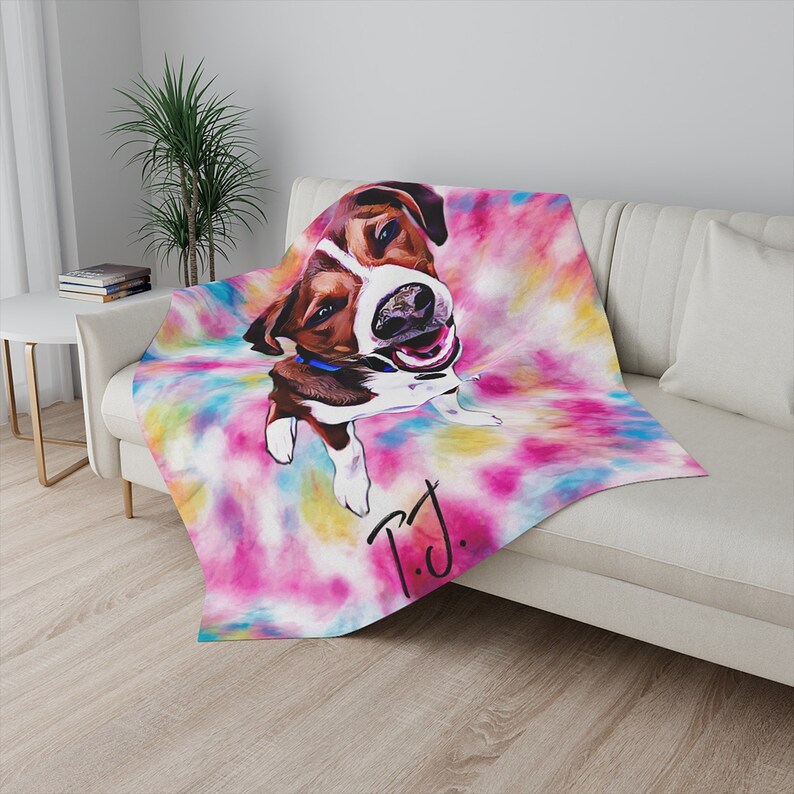 May include: A throw blanket featuring a colorful tie-dye background with a portrait of a dog. The dog is brown and white with a blue collar. The blanket has the handwritten initials "T.J." in black.