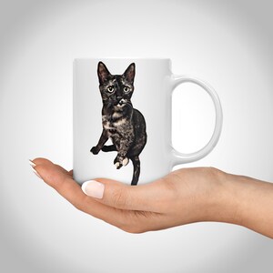 K&ouml;nnte beinhalten: Wei&szlig;e Keramiktasse mit einer Illustration einer schwarz-braunen Tabby-Katze. Die Katze sitzt mit gekreuzten Vorderpfoten und schaut in die Kamera.