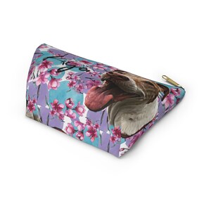 Puede incluir: Una bolsa de cosm&eacute;ticos de color morado y azul con un patr&oacute;n floral y la cara de un perro. El perro est&aacute; sacando la lengua y tiene un pelaje blanco y marr&oacute;n. La bolsa tiene una cremallera dorada.