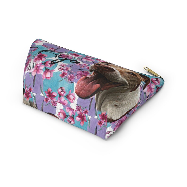 Puede incluir: Una bolsa de cosm&eacute;ticos de color morado y azul con un patr&oacute;n floral y la cara de un perro. El perro est&aacute; sacando la lengua y tiene un pelaje blanco y marr&oacute;n. La bolsa tiene una cremallera dorada.