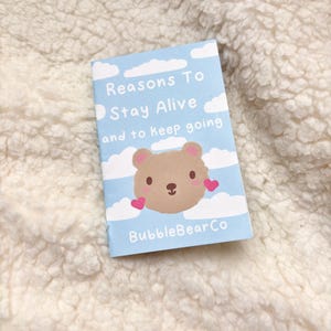 Może przedstawiać: Mała, jasnoniebieska książeczka z tekstem "Reasons To Stay Alive and to Keep Going". Okładka przedstawia kreskówkowego misia z różowymi sercami i napisem "BubbleBearCo."