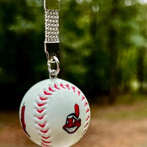 May include: A silver-toned metal keychain featuring a miniature white baseball charm with red stitching. The baseball displays a red and black graphic of a smiling cartoon character wearing a feather headdress on its side.