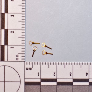May include: Small, gold-colored jewelry findings, possibly earring posts or charms, are scattered on a light blue surface. Each finding has a small loop and a straight post. The image includes a ruler for scale.