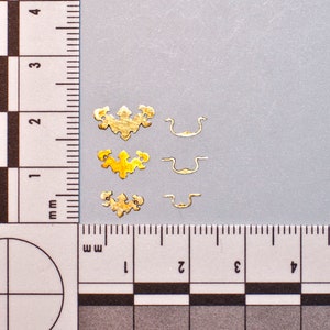 May include: A selection of small, gold-coloured decorative hardware pieces. The image shows three ornate, floral-shaped embellishments and three small, curved handle-like components. The items are arranged on a light grey surface next to a ruler.