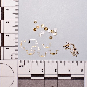 May include: A collection of small, metallic hardware components. The assortment includes gold-toned drawer pulls, rivets, and circular pieces in gold, silver, and bronze. The items are arranged on a gray surface next to a ruler with millimeter markings.