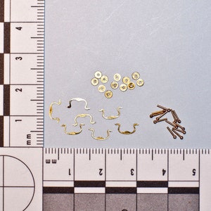 May include: A collection of small, gold-colored hardware components, including drawer pulls, round fasteners, and small pins with ball ends. The items are arranged on a gray surface next to a ruler with millimeter markings.