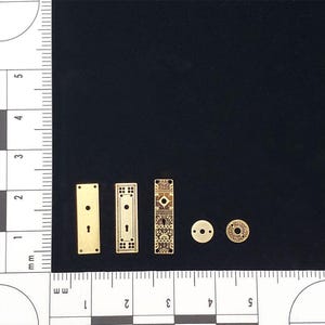 May include: A selection of miniature gold-coloured door hardware components, including rectangular plates with keyholes and decorative designs, and circular plates. The hardware is displayed on a dark surface, with a ruler for scale.