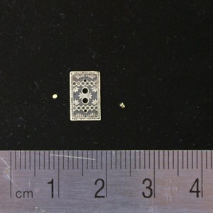 May include: A small, rectangular button with an intricate floral and geometric design. The button has two holes and is made of a light-colored material. A ruler with centimeter markings is in the foreground. The button is approximately 1 cm in length.