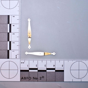 May include: Two small, decorative hinges with a cream-colored finish and gold-colored accents. Each hinge has a diamond-shaped end and a central pivot point. The hinges are displayed on a gray surface with a ruler and measurement markings.
