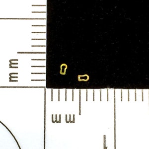 May include: Two small, gold-colored metal keyhole charms on a black background. The charms are in the shape of a keyhole and a keyhole slot. A ruler with millimeter markings is in the foreground.