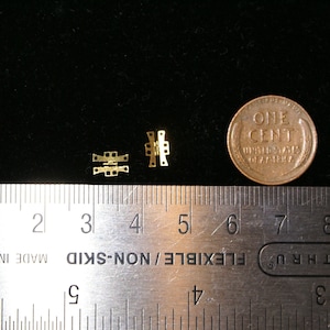 May include: Two small, gold-colored metal hinges are displayed on a black surface next to a US penny. The hinges are intricately designed with multiple rectangular and square cutouts. A ruler is in the foreground.