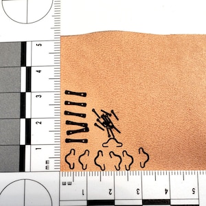 May include: An assortment of small, black metal hardware pieces, including rectangular plates with holes, small pins, and curved clasps, are arranged on a tan leather surface. A ruler with millimeter markings is in the frame.