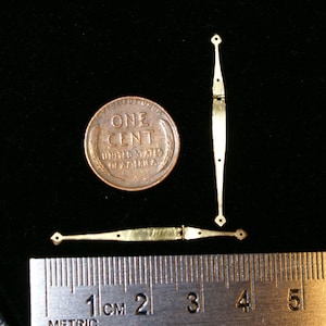 May include: Close-up of three small, gold-colored metal hinges. One U.S. penny is next to the hinges for size comparison. A ruler with centimeter markings is in the foreground. The hinges have a decorative, elongated shape with a diamond-shaped end.