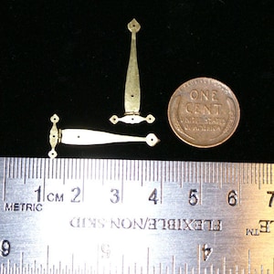 May include: Two small, brass-colored hinges with decorative finials and a penny for scale. The hinges are shown open and closed, with a ruler indicating metric measurements. The penny reads "ONE CENT UNITED STATES OF AMERICA."