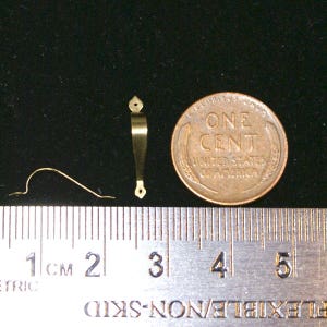 May include: A close-up of miniature gold-colored hardware, including a small handle, a curved wire, and a penny for scale. The penny reads "ONE CENT" and "UNITED STATES OF AMERICA". A ruler with centimeter markings is in the foreground.