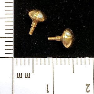 May include: Two small, gold-colored, mushroom-shaped objects with a cylindrical stem. The objects are on a black surface next to a ruler with millimeter markings. The objects have a reflective, metallic finish.